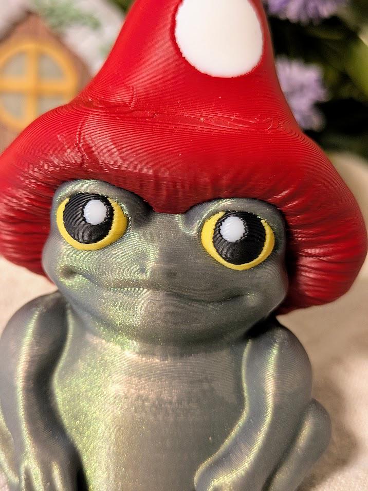 Close-up of frog figurine with wide yellow eyes and red mushroom cap, showing pearlescent texture.