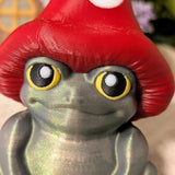 Close-up of frog figurine with wide yellow eyes and red mushroom cap, showing pearlescent texture.