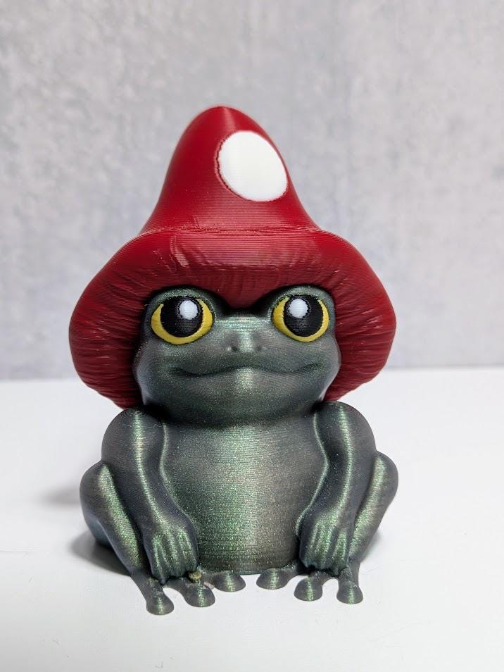 Frog figurine with shimmering green body and oversized red mushroom cap, seated against a neutral backdrop.