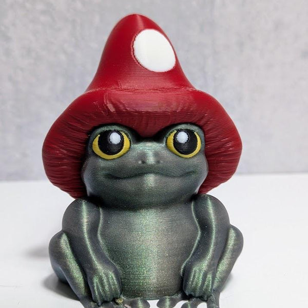 Frog figurine with shimmering green body and oversized red mushroom cap, seated against a neutral backdrop.