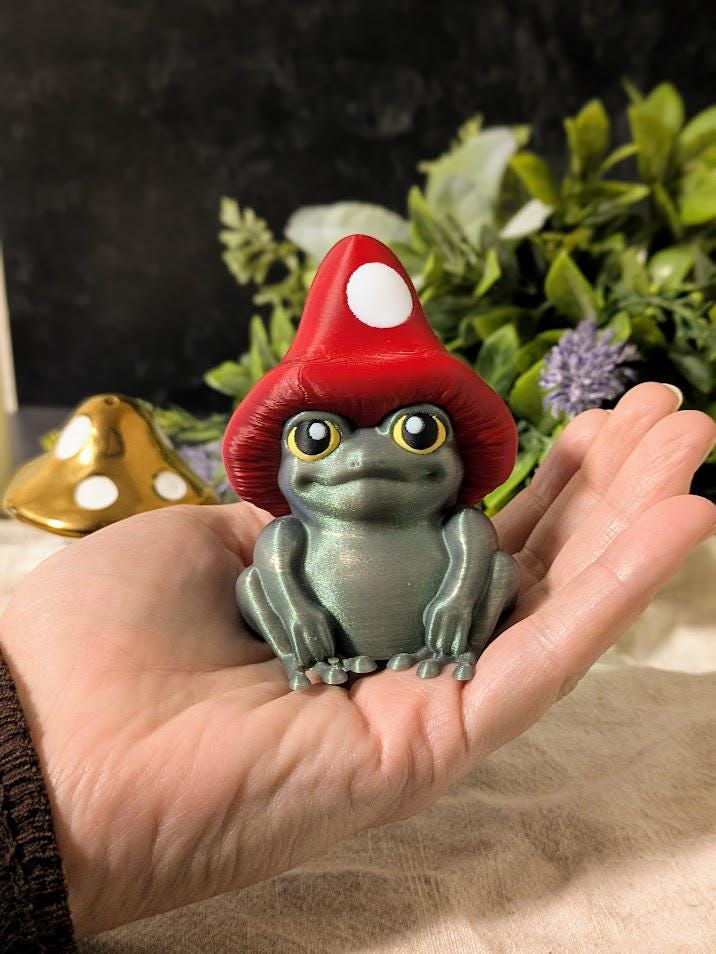 Mushroom frog figurine held in a hand for scale, with cozy warm lighting and florals in the background.