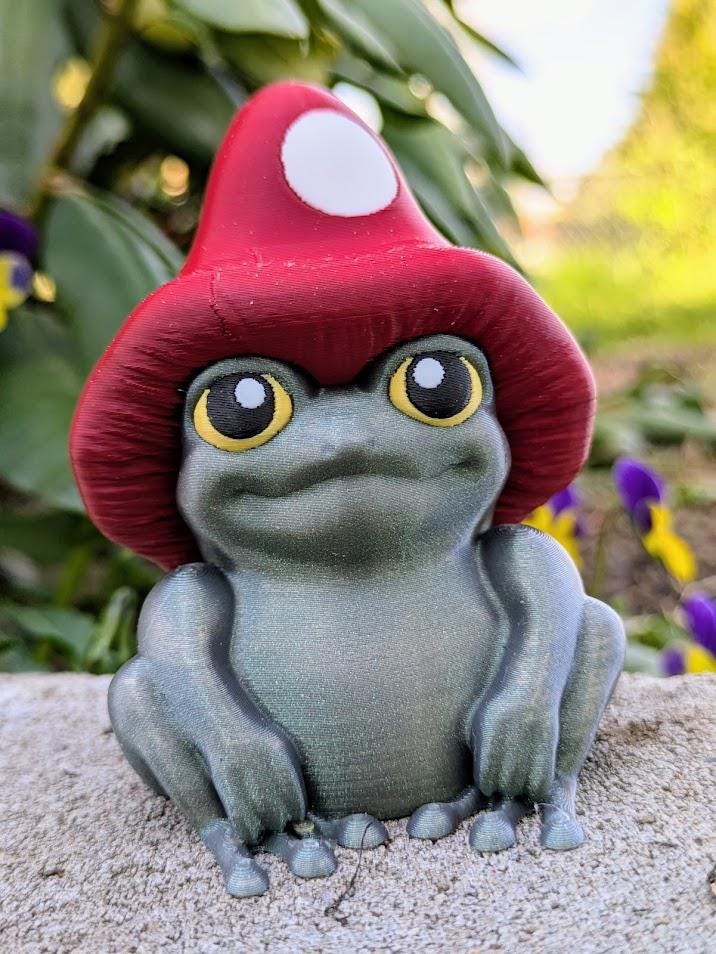 Frog figurine with red mushroom cap and yellow eyes sitting on a stone surface, with vibrant greenery in the background.