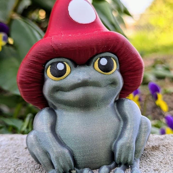 Frog figurine with red mushroom cap and yellow eyes sitting on a stone surface, with vibrant greenery in the background.