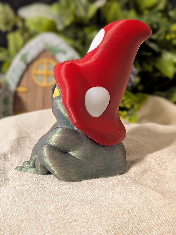 Side view of frog figurine wearing a red mushroom cap, sitting on cloth with miniature fairy door in background.