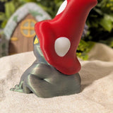 Side view of frog figurine wearing a red mushroom cap, sitting on cloth with miniature fairy door in background.