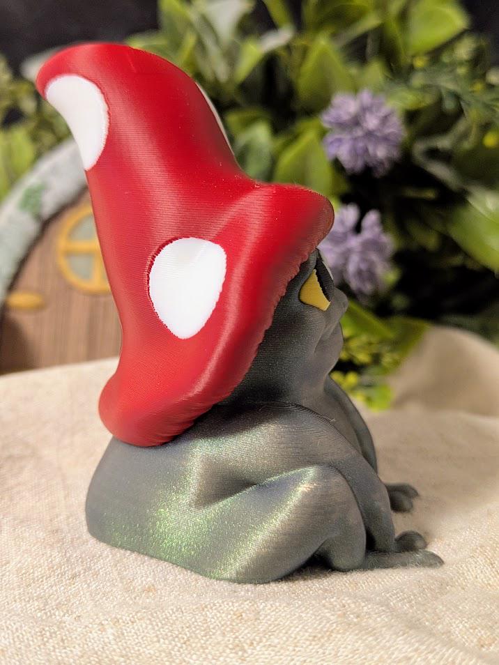 Alternate side view of frog in mushroom cap facing right, surrounded by soft florals and miniature scenery.