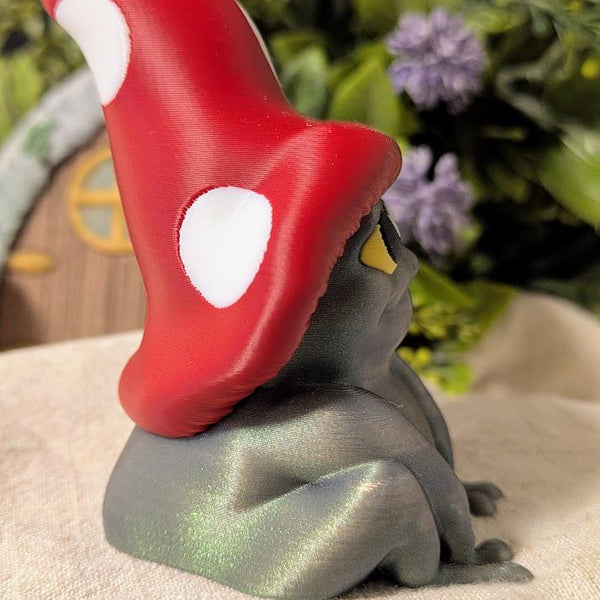 Alternate side view of frog in mushroom cap facing right, surrounded by soft florals and miniature scenery.