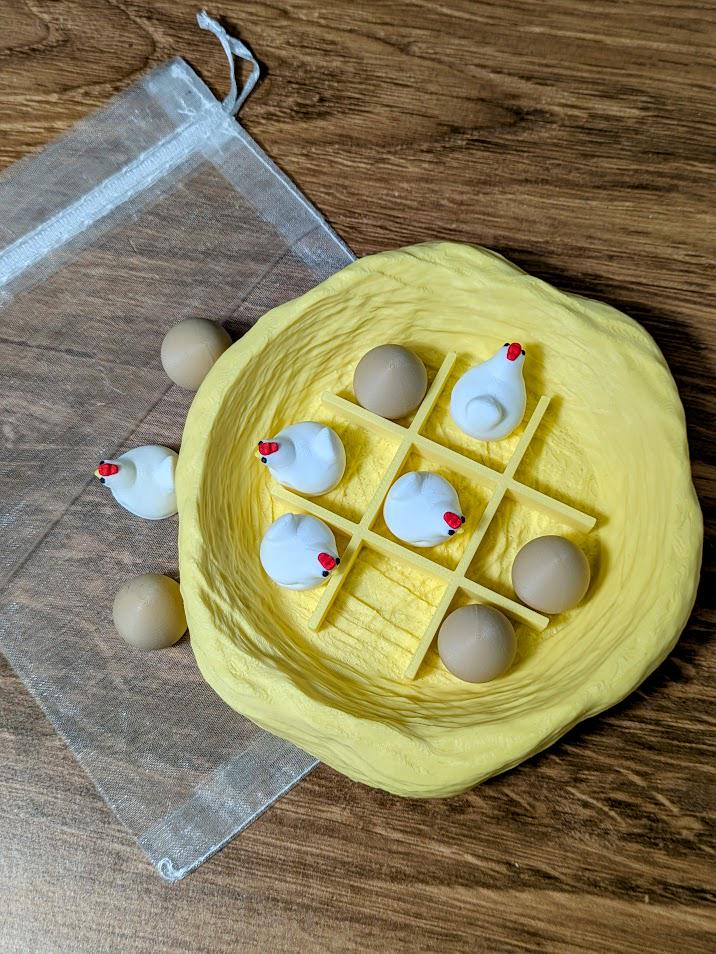 Yellow nest-shaped tic tac toe board filled with miniature chicken and egg pieces, next to an organza bag.