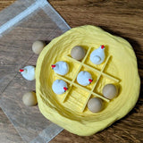 Yellow nest-shaped tic tac toe board filled with miniature chicken and egg pieces, next to an organza bag.