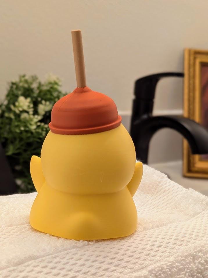 Back view of plunger hat duck figurine, posed in a bathroom scene with faucet in the background.