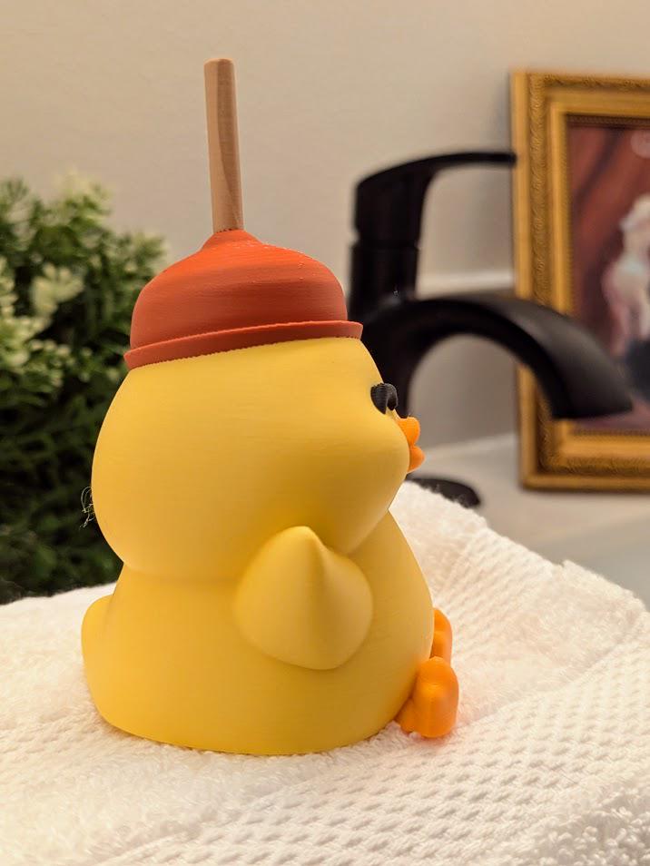 Angled side-back view of plunger hat duck figurine, perched on a towel with bathroom decor in the background.