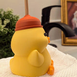 Angled side-back view of plunger hat duck figurine, perched on a towel with bathroom decor in the background.