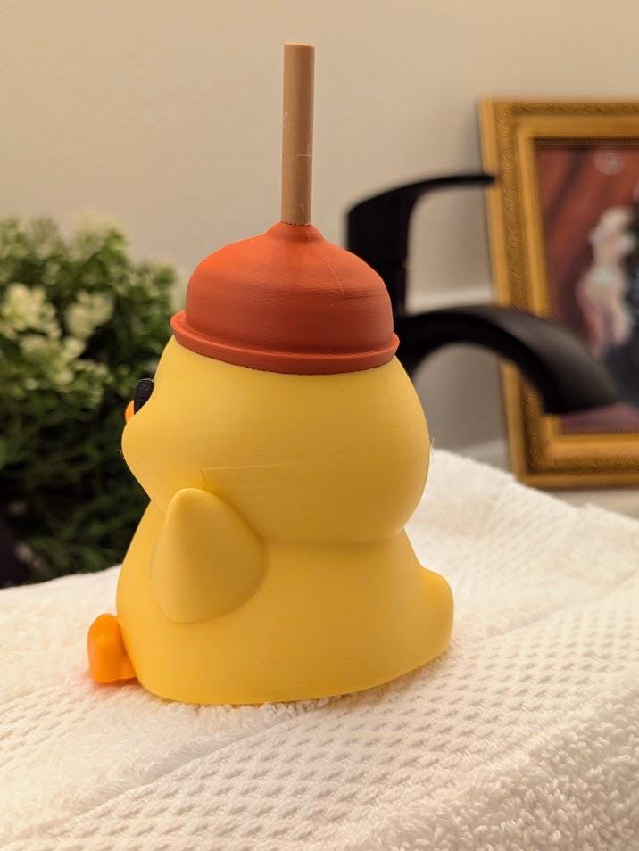 Side view of duck figurine wearing a red plunger hat, perched on a towel in front of a bathroom faucet.