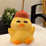 Smiling yellow duck figurine wearing a red plunger as a hat, sitting on a white towel in a bathroom setting.