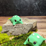 A small green blob frog figurine is shown from multiple angles, gently held in hand. The soft curves, big eyes, and mushroom-like spots give it an adorably squishy appearance as it rests on moss and stone. Its expression remains blank yet deeply relatable.