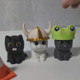 Three whimsical cat figurines in interchangeable hats, including a frog hat, cheese hat, and witch hat.