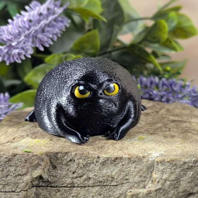 Front-facing black rainfrog figurine with grumpy yellow eyes and stubby limbs, perched on a stone among purple flowers.