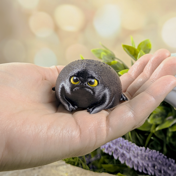 A grumpy black rainfrog figurine with yellow eyes sits in the palm of a hand, showing its small size and chonky, textured body.
