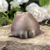 Back view of a wide, metallic green frog figurine sitting on a stone surface, showing only its rounded body and stubby legs.