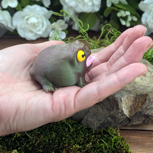 Side view of the Screaming Frog held in a hand, showing its shiny green body, yellow eyes, and exaggerated open mouth.