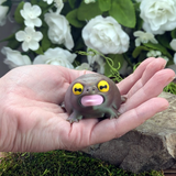 Front view of the Screaming Frog figurine resting in a hand, mouth open wide with a pink tongue and bulging yellow eyes.