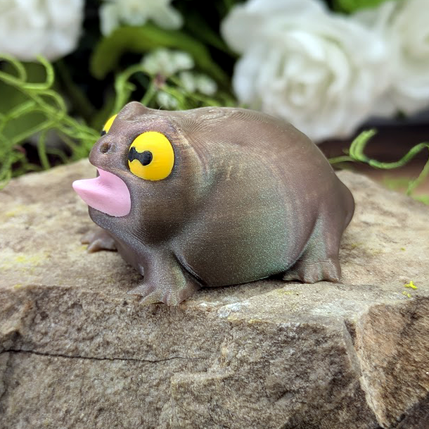 Three-quarter left angle of the frog figurine sitting on a stone, mouth open with a curved pink tongue and dramatic expression.