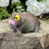 Three-quarter left angle of the frog figurine sitting on a stone, mouth open with a curved pink tongue and dramatic expression.