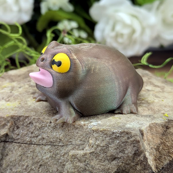 Three-quarter left angle of the frog figurine sitting on a stone, mouth open with a curved pink tongue and dramatic expression.