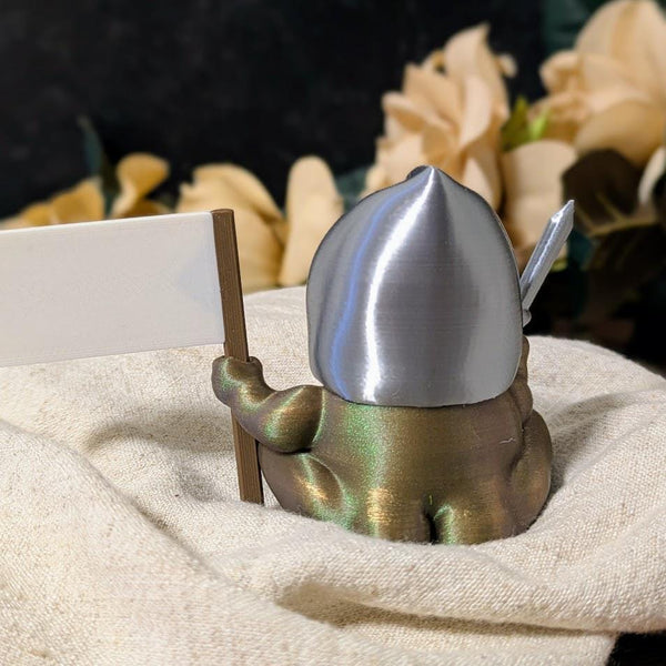 Rear view of Sir Glorb™ the frog knight holding a flag, sitting on a soft surface with floral backdrop.