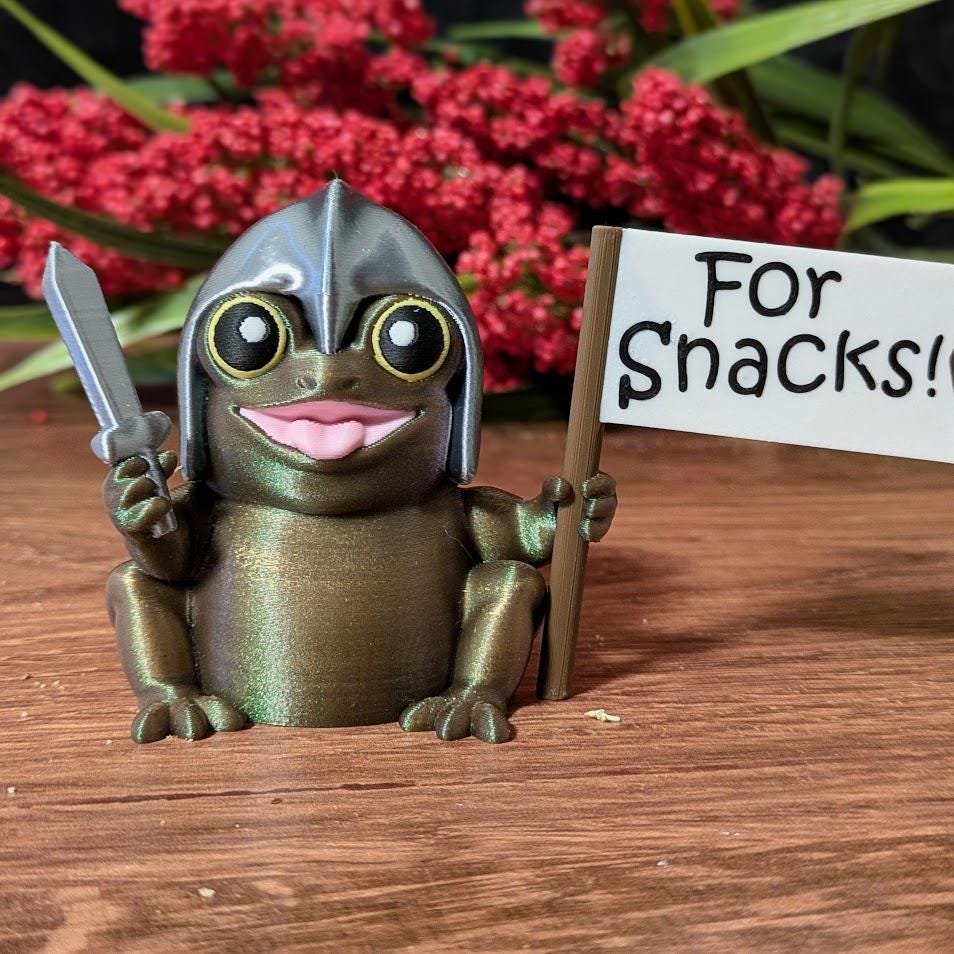 Sir Glorb™ figurine with sword and “For Snacks!” flag posed in front of red flowers on wooden surface.
