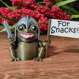 Sir Glorb™ figurine with sword and “For Snacks!” flag posed in front of red flowers on wooden surface.