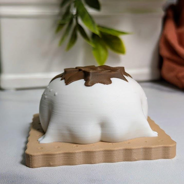 A marshmallowy frog figurine with chocolate drips on its back sits atop a textured graham cracker base, facing backwards with tiny frog feet peeking out.