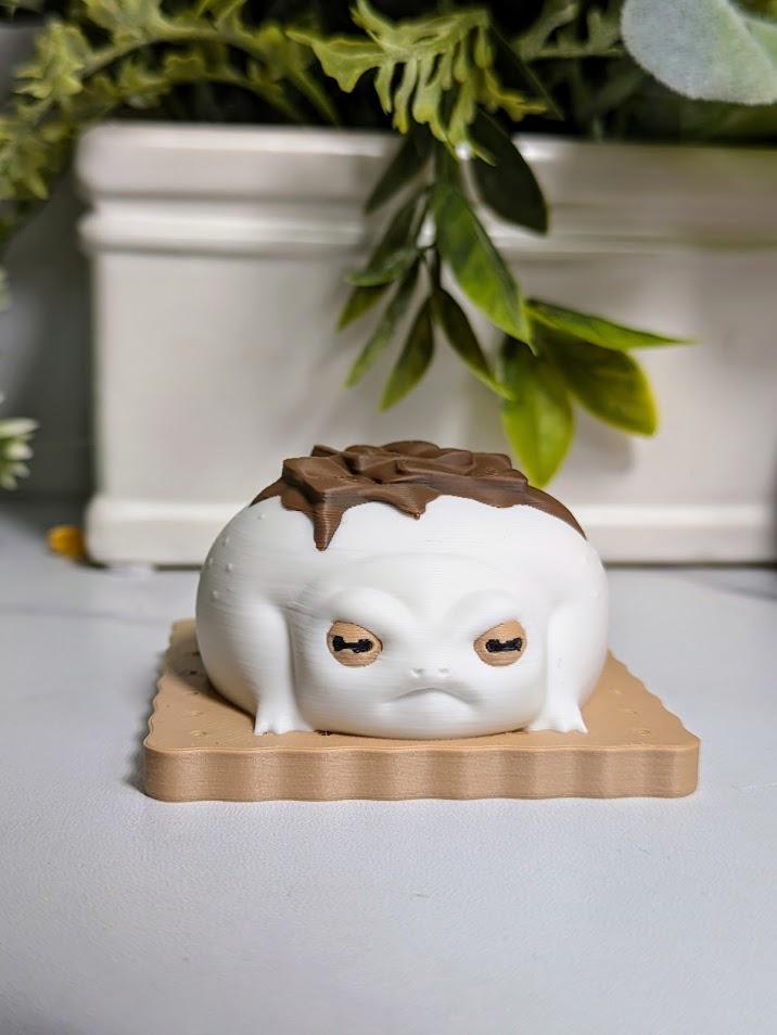 Front-facing close-up of a frog figurine styled like a s’more, with big eyes and a chocolate accent on its back, sitting neatly on a graham base.