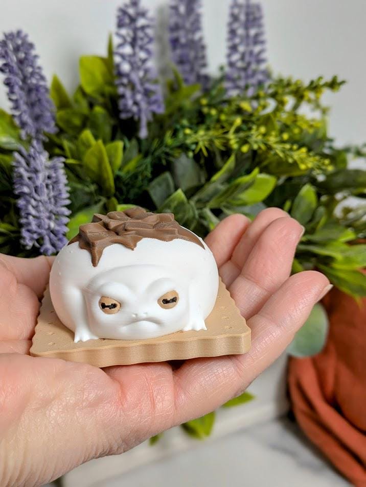 A person holds a small s’mores frog figurine in their palm. The frog has a marshmallow-white body and a melty chocolate top, with plants in the background.