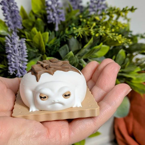 A person holds a small s’mores frog figurine in their palm. The frog has a marshmallow-white body and a melty chocolate top, with plants in the background.