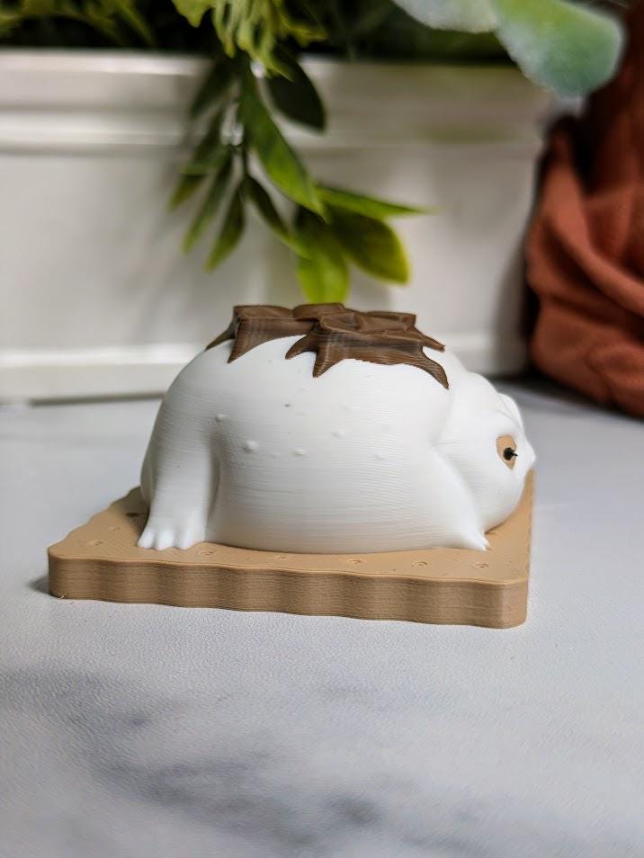 Side profile of a s’mores-inspired frog, showing its soft white body and melted chocolate detail across its back while sitting on a graham cracker.