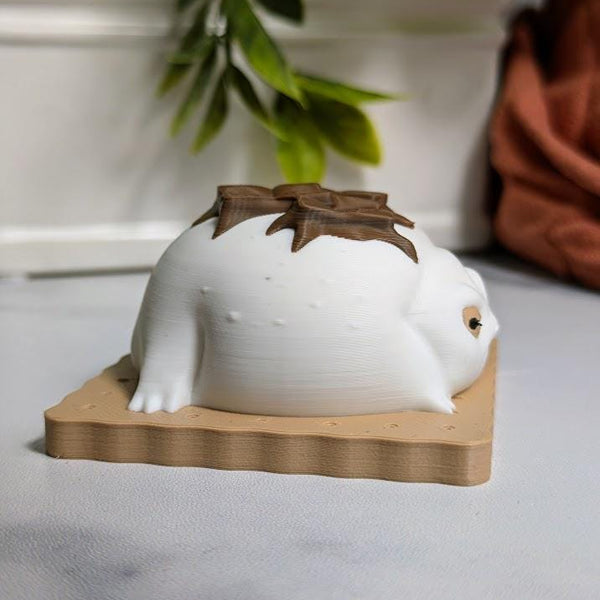 Side profile of a s’mores-inspired frog, showing its soft white body and melted chocolate detail across its back while sitting on a graham cracker.