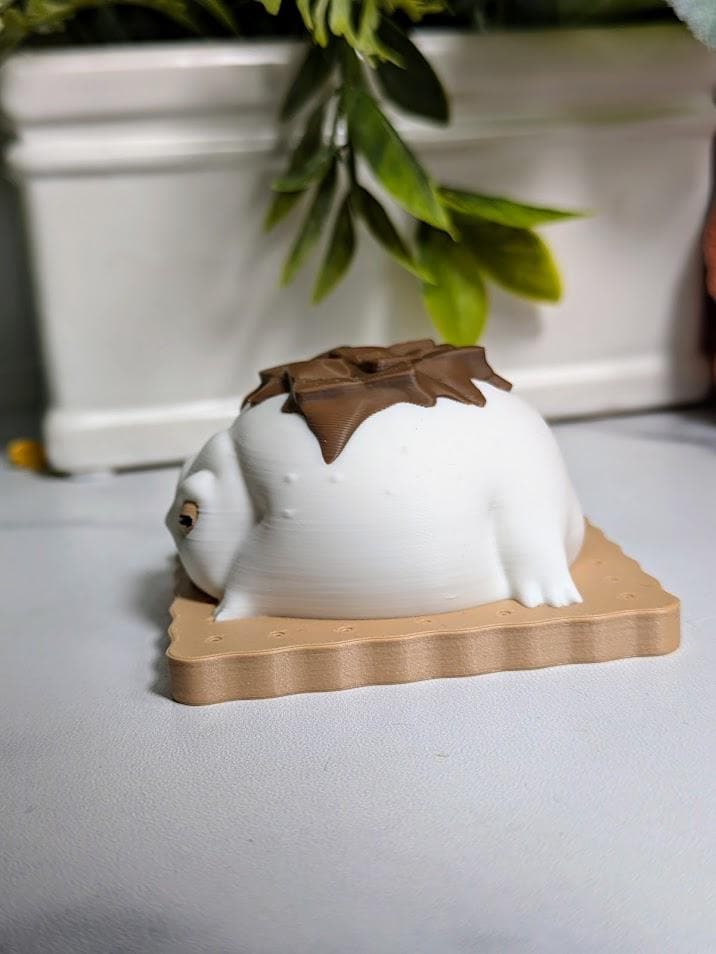 Side view of a s’mores-inspired frog figurine, showing a smooth marshmallow body, pink feet, and drippy chocolate shell on a cracker.