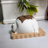 Side view of a s’mores-inspired frog figurine, showing a smooth marshmallow body, pink feet, and drippy chocolate shell on a cracker.
