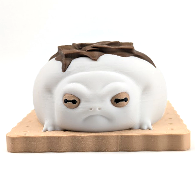 A very grumpy looking white frog marshmallow on top of a graham cracker and topped with chocolate. This smores frog sits on a white backdrop.