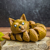Front view of the Snat figurine curled on a stone surface, showing its cat face and scaled snake body in shimmering golden PLA.