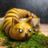 Close-up of a 3D printed snake-cat hybrid figurine with an intense, slightly angry cat face and a glistening golden brown body, posed on green moss.