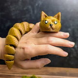 The articulated Snat figurine wrapped loosely around a hand, displaying its long flexible snake body and expressive feline head.