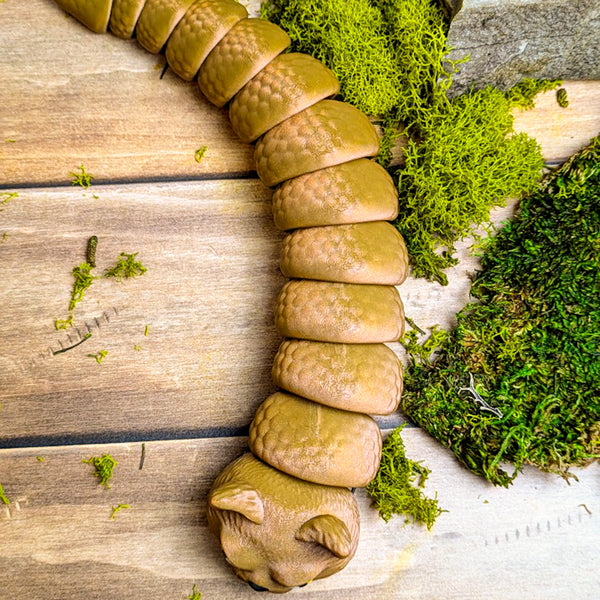 Top-down view of the Snat figurine fully extended on a wooden surface, with its sparkly brown scales and cat head resting near green moss.