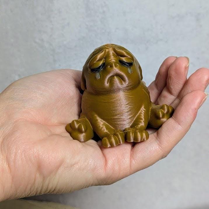 Metallic bronze "sad potato brown" Squonk figurine sitting in a person’s palm, weeping quietly into the void.