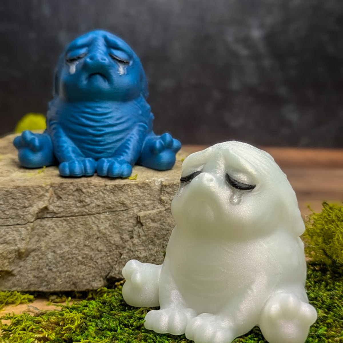 A Moonstone Tears Squonk sitting on moss and crying, with a sobbing Cerulean Sadness squonk on a rock in the background.