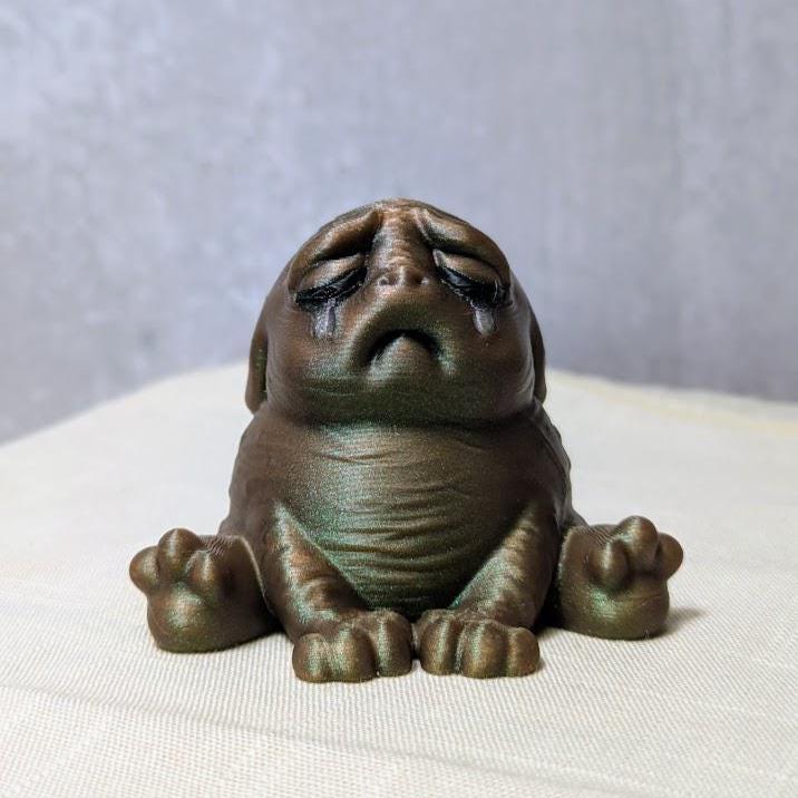 Swamp Grief Green Squonk sitting upright on a pale surface, visibly crushed by the weight of existence.