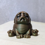 Swamp Grief Green Squonk sitting upright on a pale surface, visibly crushed by the weight of existence.