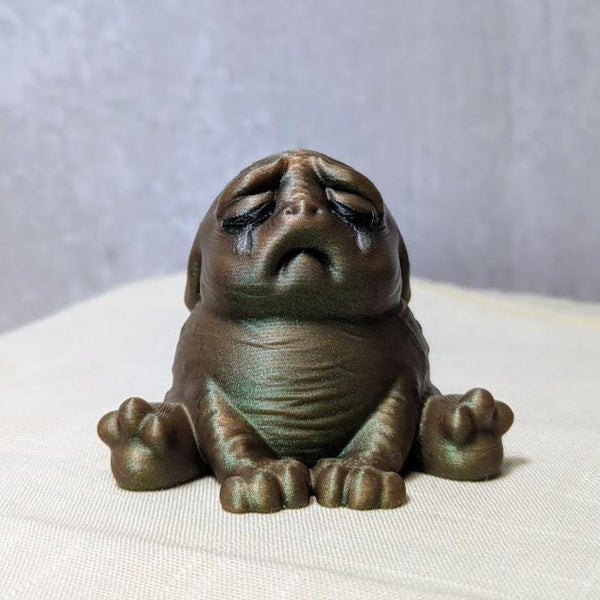 Swamp Grief Green Squonk sitting upright on a pale surface, visibly crushed by the weight of existence.