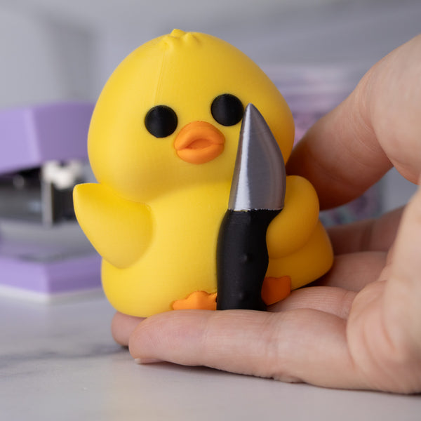 Yellow duck holding a knife with a blurred background, held in a hand for scale.